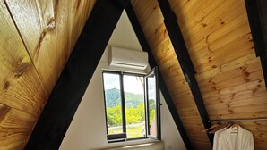 A bedroom with a wooden sloped ceiling, a bed with white linens, a brown throw blanket, and a window with a view of greenery.