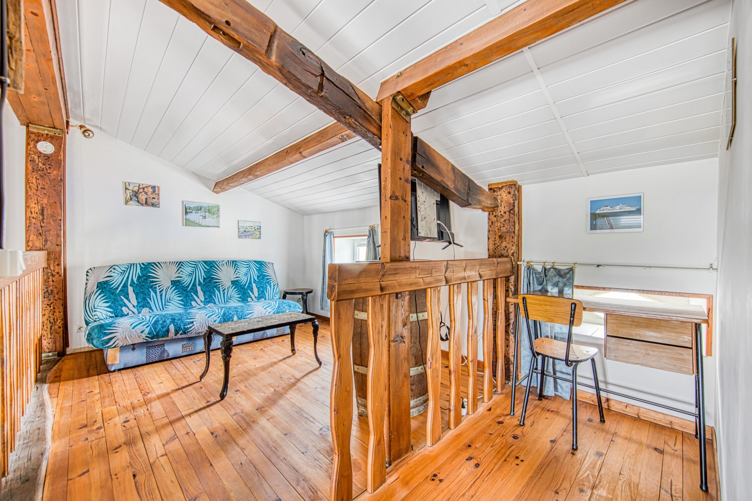 A room with wooden beams, a sofa with a leaf pattern, a desk with a chair, and a window with a view.