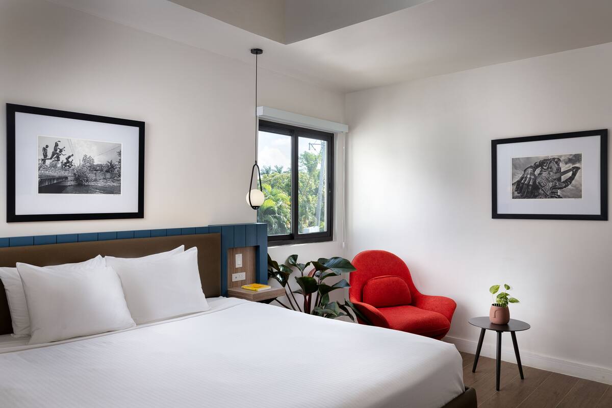 Une chambre d’hôtel moderne avec un grand lit, un fauteuil rouge, une petite table avec une plante et deux tableaux encadrés accrochés aux murs.