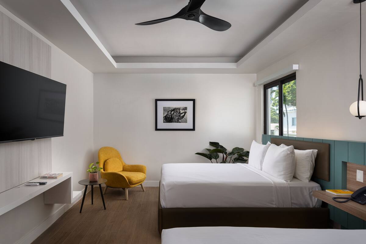 Une chambre d’hôtel moderne équipée d’une télévision à écran plat, d’un fauteuil jaune, d’un lit avec du linge de maison blanc et d’un ventilateur de plafond.
