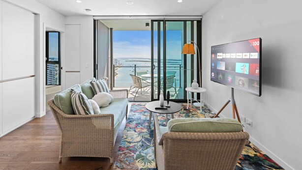 A modern living room with a large TV, wicker furniture, a colorful rug, and a balcony with a view of the sea. - CASSA MQ - Oceanview 2BR Southport Gem for Family (Southport)
