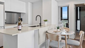 A modern kitchen with white cabinetry, a central island with a sink, and a dining area with a round table and chairs. - CASSA MQ - Oceanview 2BR Southport Gem for Family (Southport)