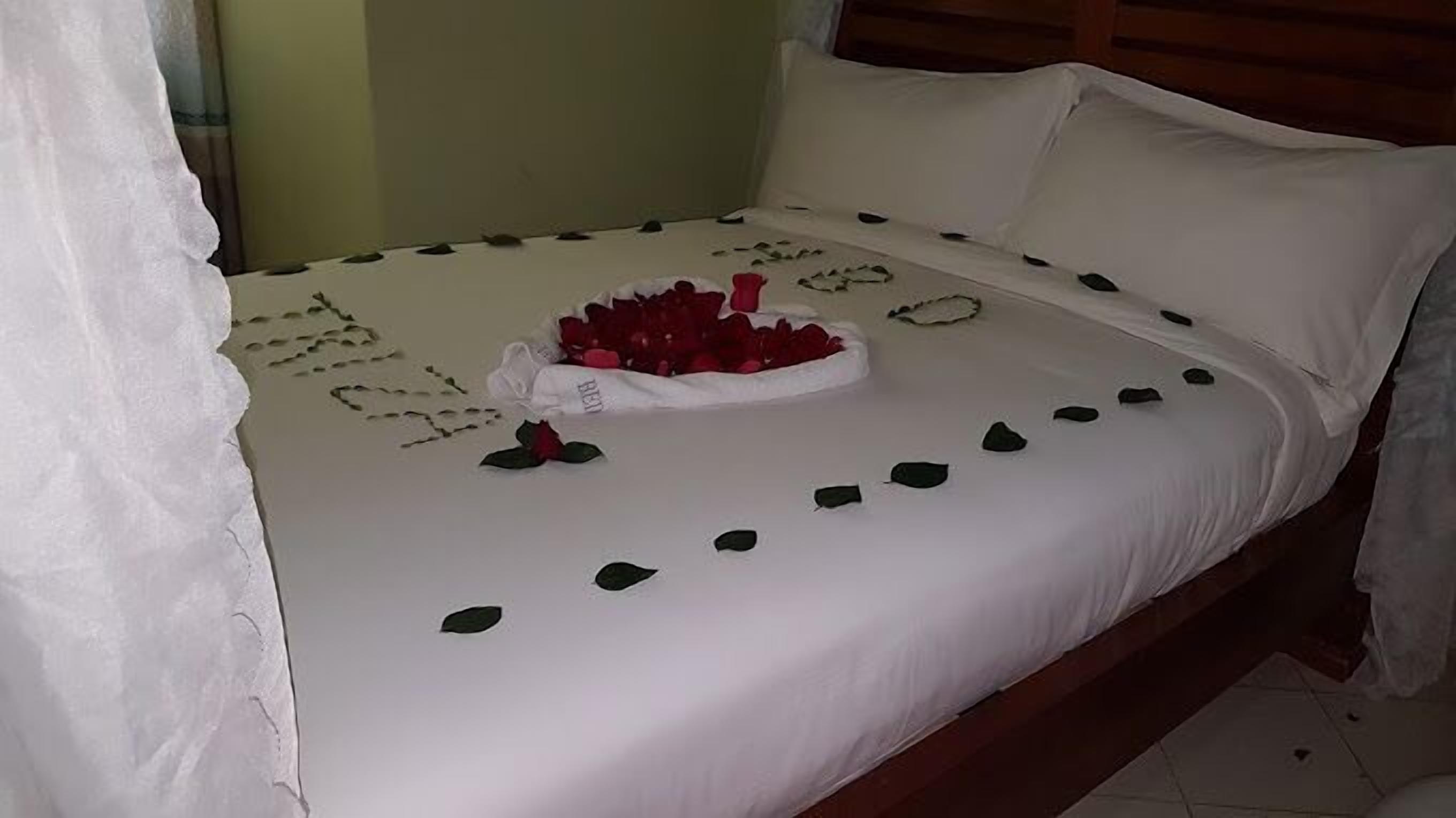 A bed with a white bedspread, a decorative arrangement of rose petals, and a small bouquet of red roses.