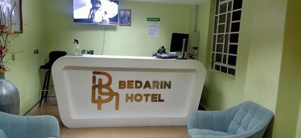 A reception desk with a logo for Bedarin Hotel, a television screen, and a window with blinds. - Bedarin Hotel Ruiru (Ruiru)