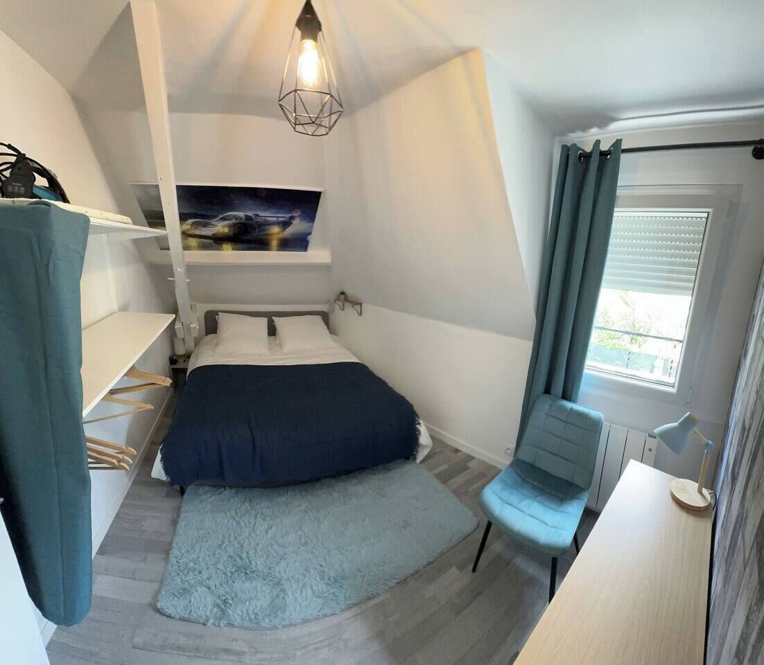 Un dormitorio pequeño con cama, escritorio, silla, lámpara colgante y ventana con cortinas.