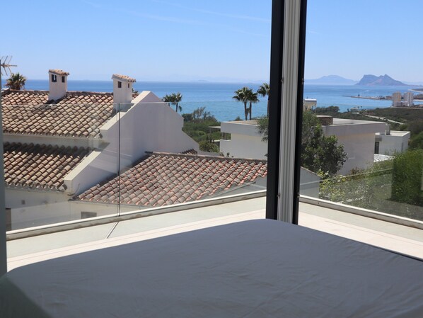A room with a large window overlooking a coastal area, featuring a tiled roof, a bed, and a view of the sea and distant mountains.