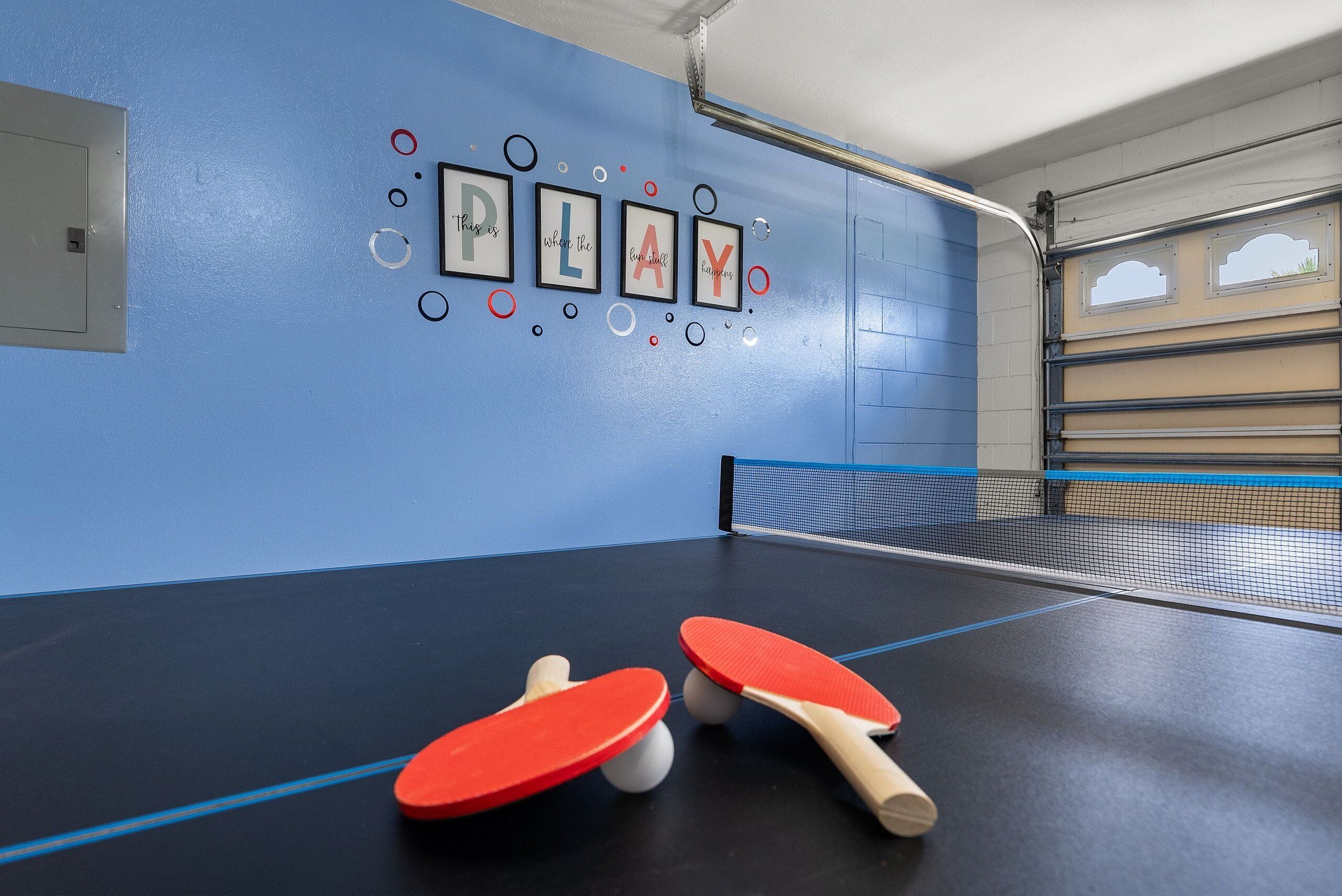 Une table de ping-pong avec deux raquettes rouges et des balles, un mur bleu orné de citations motivantes encadrées, et une porte avec une poignée en métal.