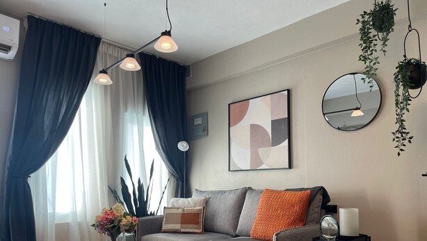 Una sala de estar moderna con un sofá gris, una mesa redonda de centro y una mesa con cubierta de vidrio y flores. Se observa un ventanal con cortinas azules, un espejo fijado a la pared y un cuadro abstracto enmarcado.
