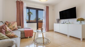 A modern living room with a sofa, a small round table, a TV, and a view of palm trees through a window.