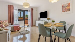 A modern living room with a glass dining table, cushioned chairs, a sofa, a TV, and a painting on the wall.