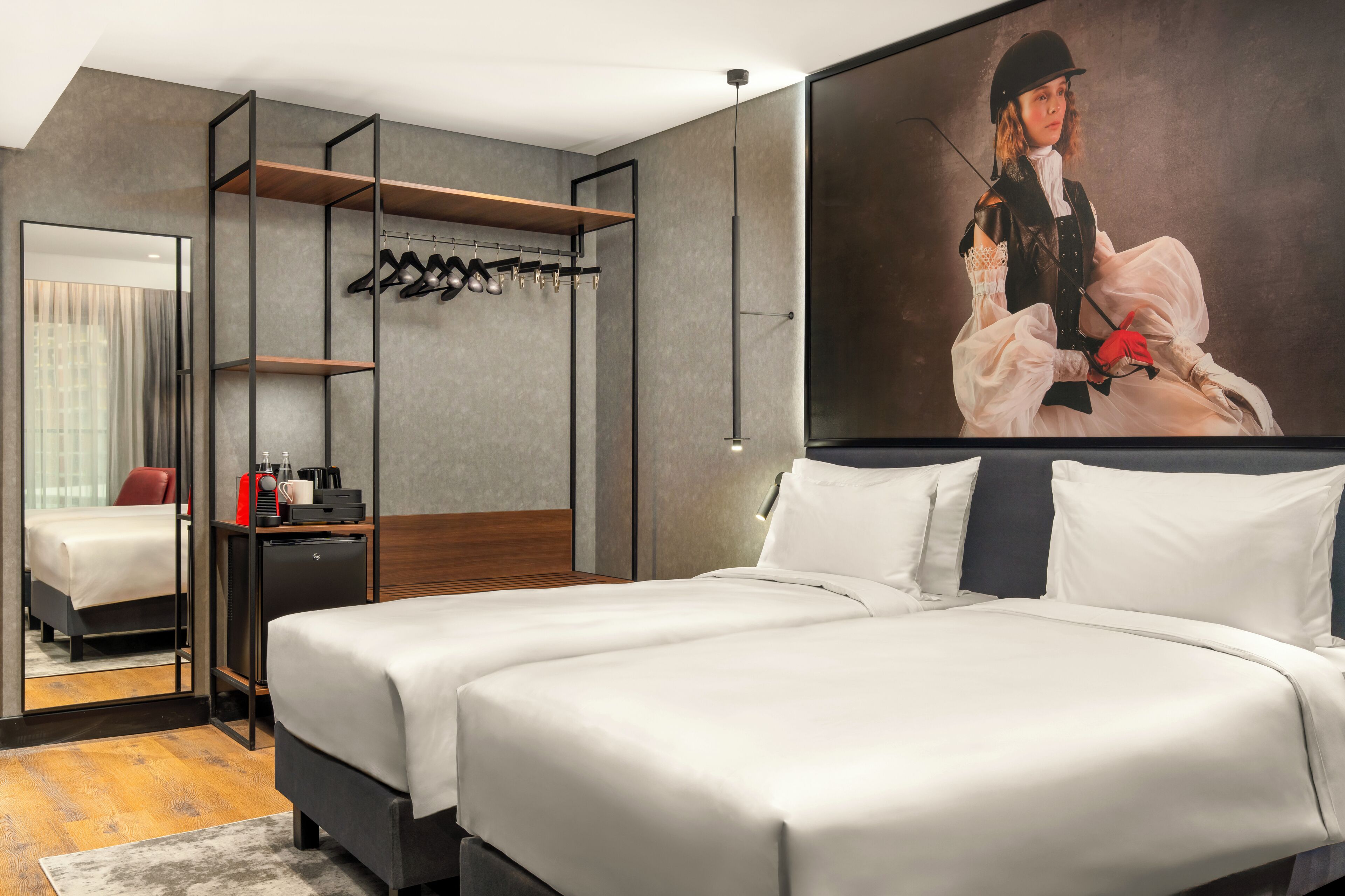 A modern hotel room with a large bed, a mirror, a coat rack, and a painting of a person in historical attire.