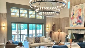 A spacious living room with a high ceiling, wooden beams, a large chandelier, a fireplace, and a large window offering a view of the outdoors. - Luxury Barn Retreat, Mountain Views and Creekside Calm (Ellijay)