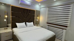 A modern bedroom with a large bed, a headboard with geometric patterns, bedside tables, and a ceiling with blue and yellow lighting. - YASMİNE SUİTE (Trabzon)