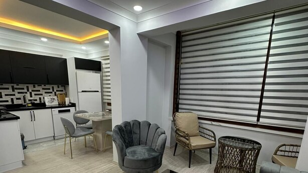 A modern living room with a sofa, armchair, and coffee table, a kitchen area, and a large window with blinds. - YASMİNE SUİTE (Trabzon)