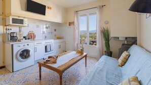 A compact living space with a kitchen, a sofa, a table, and a window with a view. - Gîte 'Canigou' with Mountain View, Wi-Fi, and Air Conditioning (Banyuls-dels-Aspres)