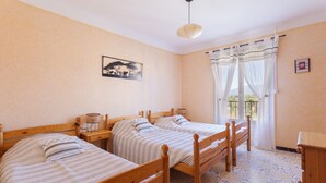 A room with two wooden beds, a small table, a hanging light fixture, and a window with curtains. - Gîte 'Canigou' with Mountain View, Wi-Fi, and Air Conditioning (Banyuls-dels-Aspres)