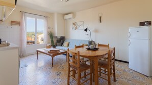 A compact kitchen with a dining table, chairs, a refrigerator, and a window with curtains. - Gîte 'Canigou' with Mountain View, Wi-Fi, and Air Conditioning (Banyuls-dels-Aspres)