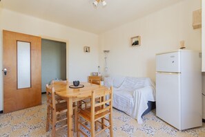 A small, cozy room with a wooden dining table, a white sofa, a white refrigerator, and a wooden door.