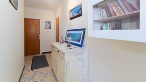 Un couloir avec une porte en bois, un tableau encadré accroché sur un meuble blanc, une bibliothèque remplie de livres et un sol carrelé.