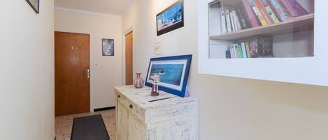 A hallway with a wooden door, a framed picture on a white cabinet, a bookshelf with books, and a tiled floor.