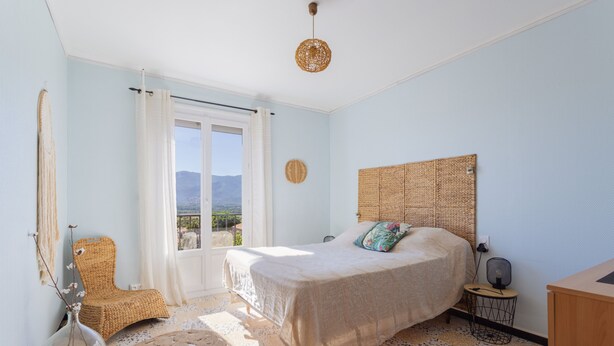 Une chambre à coucher avec un lit, une chaise, une petite table, un ventilateur et une vue sur l’extérieur.
