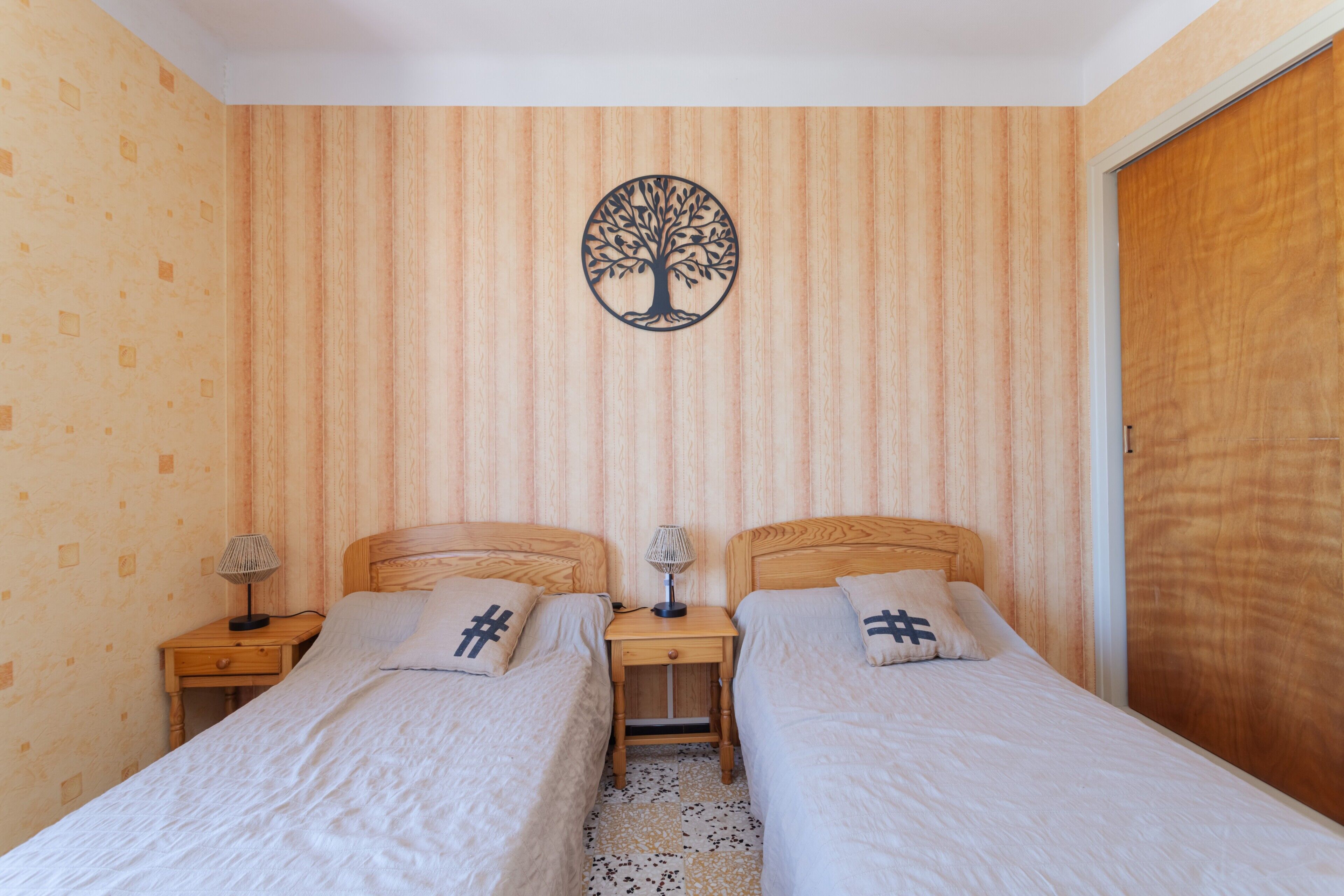 A bedroom with two beds, a nightstand with lamps, a wall-mounted tree decoration, and a wooden wardrobe.