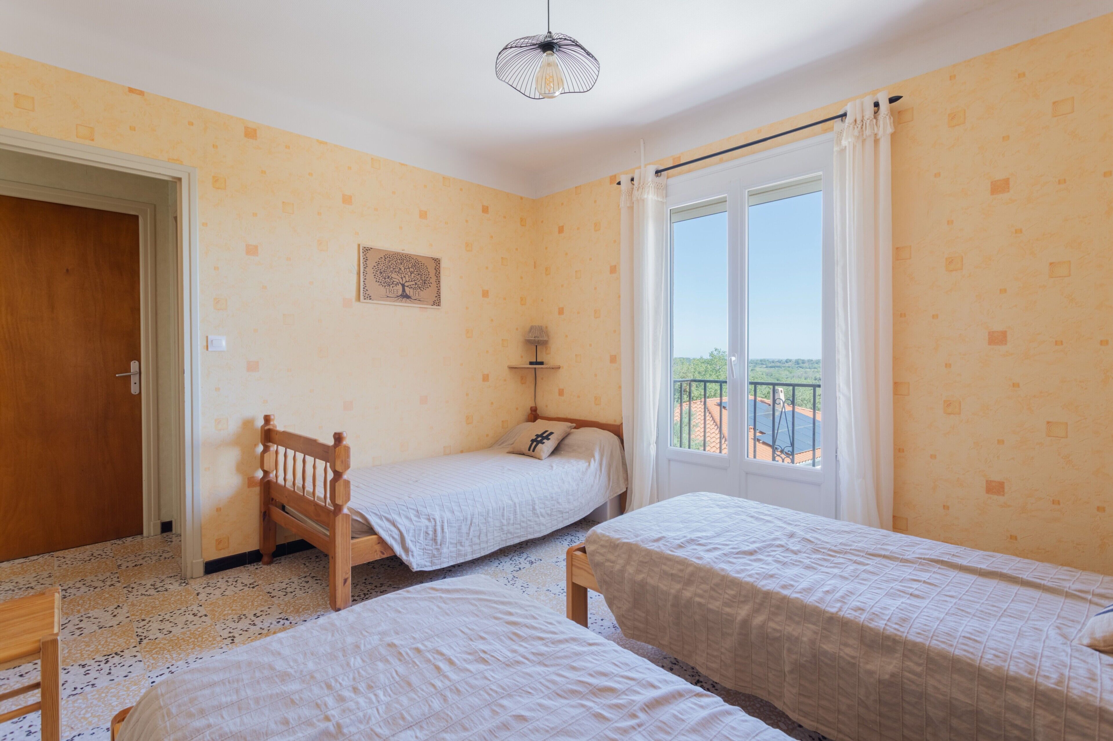 A room with two beds, a door, a window with curtains, a hanging light fixture, and a small table.