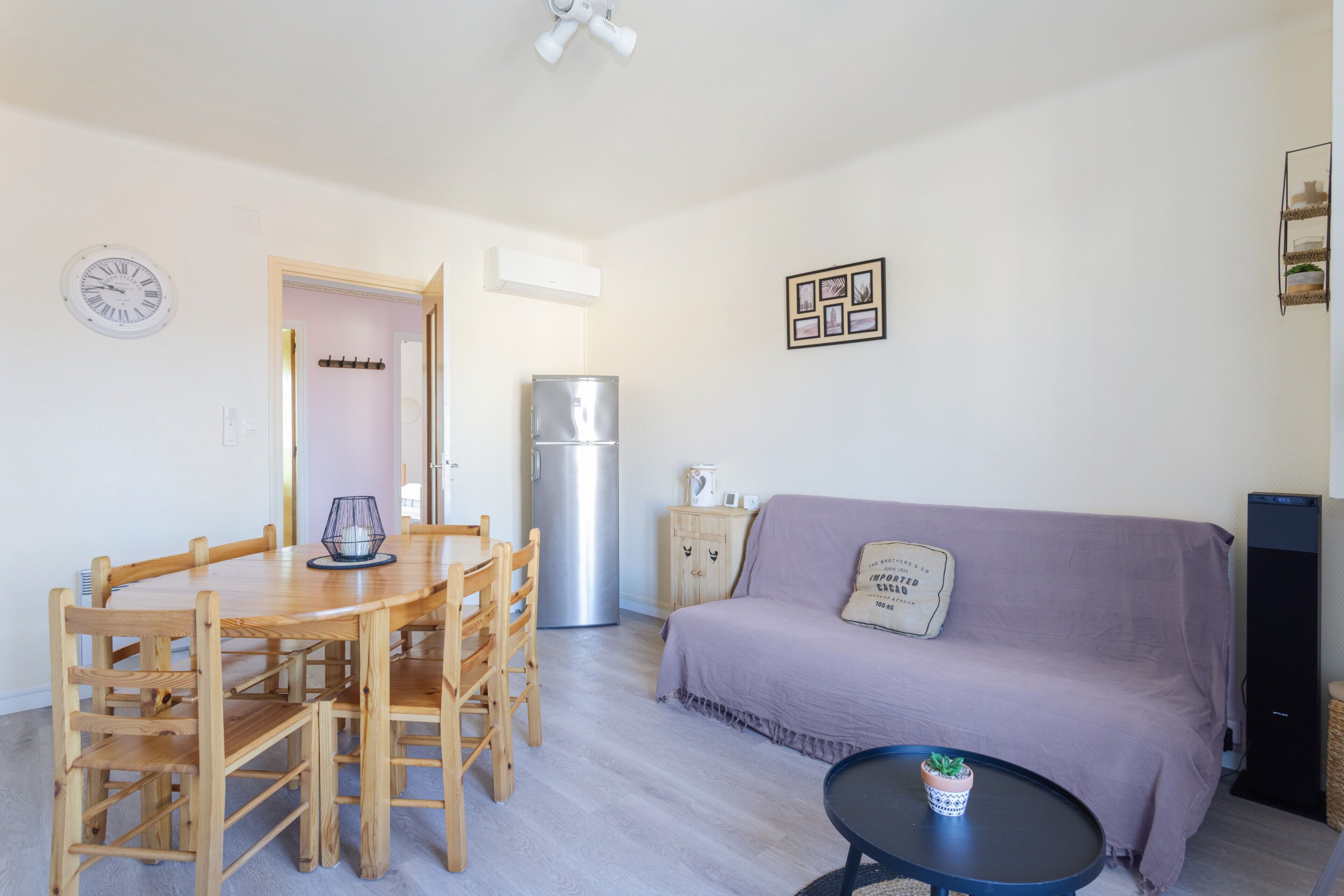 A living room with a sofa, a dining table with chairs, a small table with a plant, a refrigerator, and a clock on the wall.