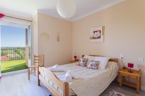 A bedroom with a bed, a chair, a nightstand, a wall clock, a dreamcatcher, and a window with a view of a green landscape.