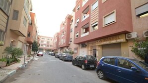 Une rue étroite en milieu urbain, bordée d’immeubles résidentiels à plusieurs étages, de voitures garées et d’une devanture de magasin bien visible avec une enseigne.