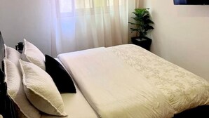 Un lit bien fait, avec une couette à motifs floraux, une plante en pot et un téléviseur fixé au mur.
