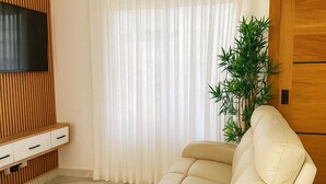 Una sala moderna con un sofá beige, un televisor de pantalla plana montado en un panel de madera, un ventanal con cortinas translúcidas y una puerta de madera.