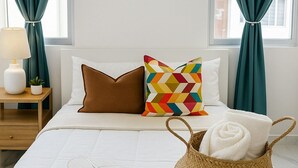 Una cama bien tendida con ropa blanca, una almohada marrón y una almohada geométrica colorida, además de una cesta tejida con toallas enrolladas.
