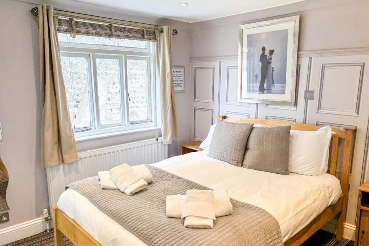A neatly made bed with a beige quilt and pillows, a window with curtains, and a framed picture on the wall.