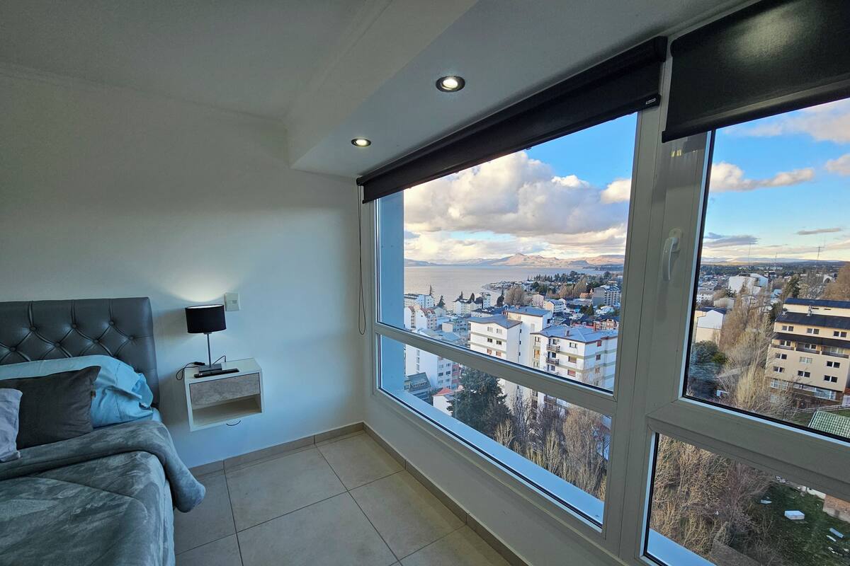 A bedroom with a large window, a bed, a bedside table, and a lamp.