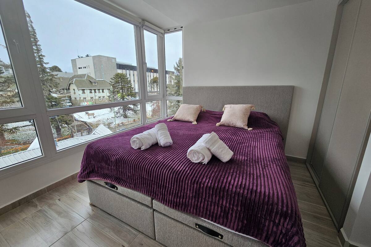 A bedroom with a large window, a bed with a purple blanket, two pillows, and rolled towels.