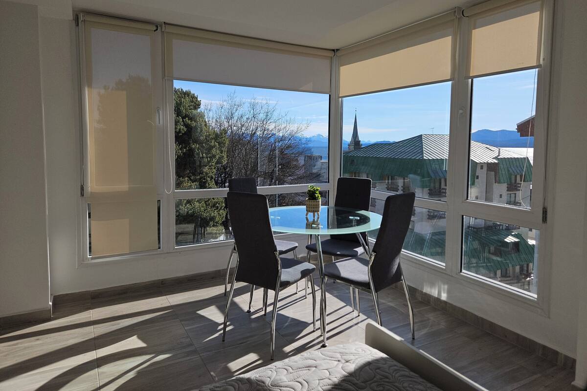A room with a glass table and four chairs, large windows with a view of a church and mountains, and a patterned rug on the floor.