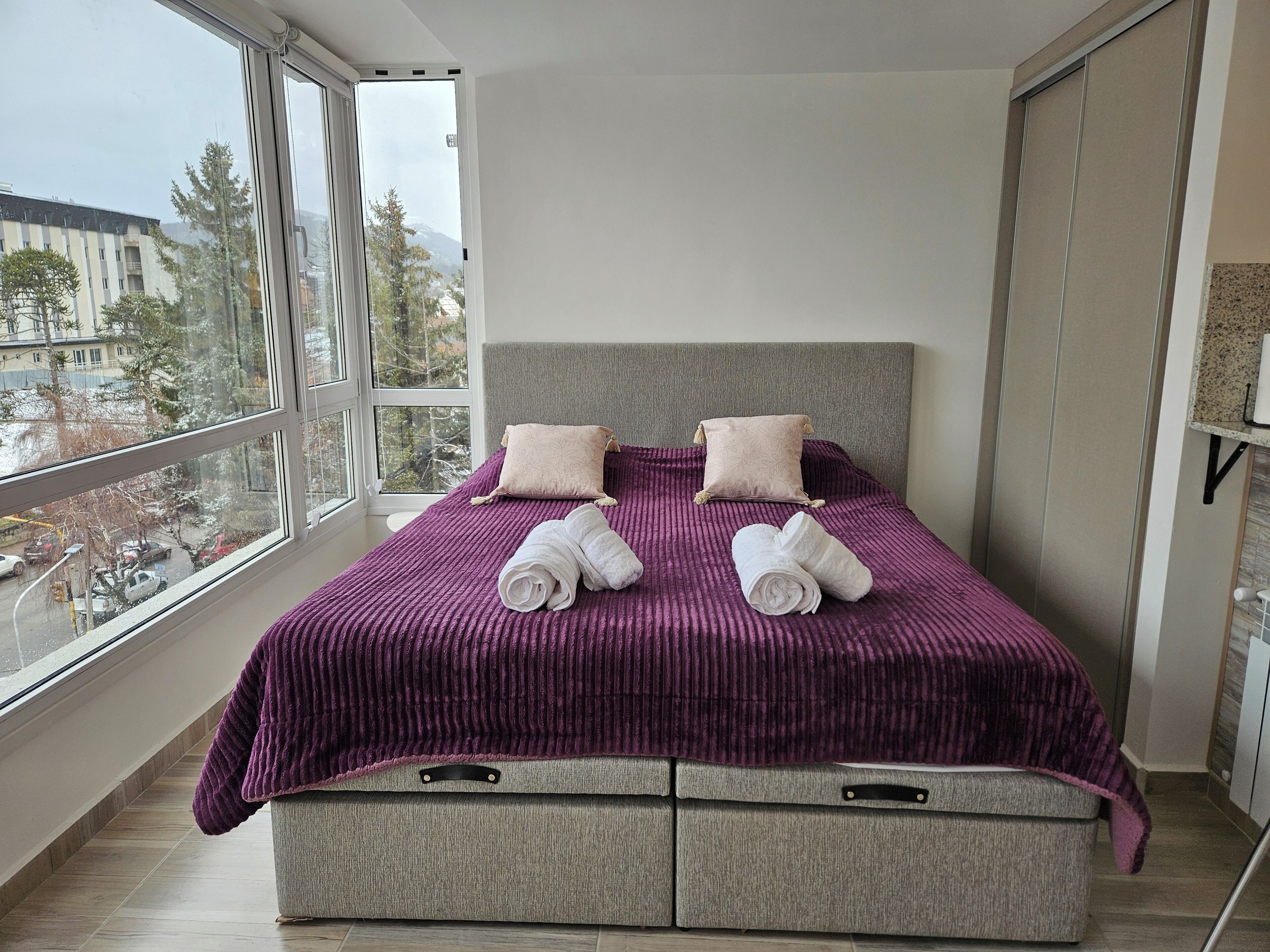 Una camera da letto moderna con una grande finestra, un letto con copriletto viola, due cuscini e due asciugamani arrotolati.