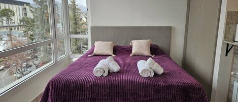 Una camera da letto moderna con una grande finestra, un letto con copriletto viola, due cuscini e due asciugamani arrotolati.