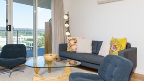 A modern living room with a glass coffee table, two armchairs, a sofa with colorful pillows, a floor lamp, and a large window with a view of buildings and trees.
