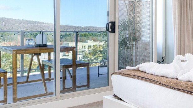A balcony with a table and chairs, a bed with a white comforter, and a view of a cityscape and hills.