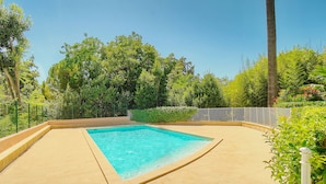 Una piscina rectangular con solárium de baldosas, rodeada de vegetación exuberante y una cerca de metal.