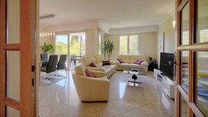 Un salón amplio con un gran sofá, una mesa de centro y un televisor de pantalla plana. Varias ventanas ofrecen luz natural y vistas al exterior.