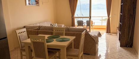 A living room with a sofa, dining table, chairs, and a view of the sea through a large window.