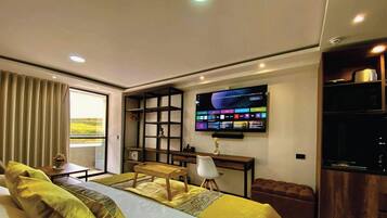 A modern hotel room with a large bed, a desk with a computer, a TV mounted on the wall, and a view of the outdoors through a window.