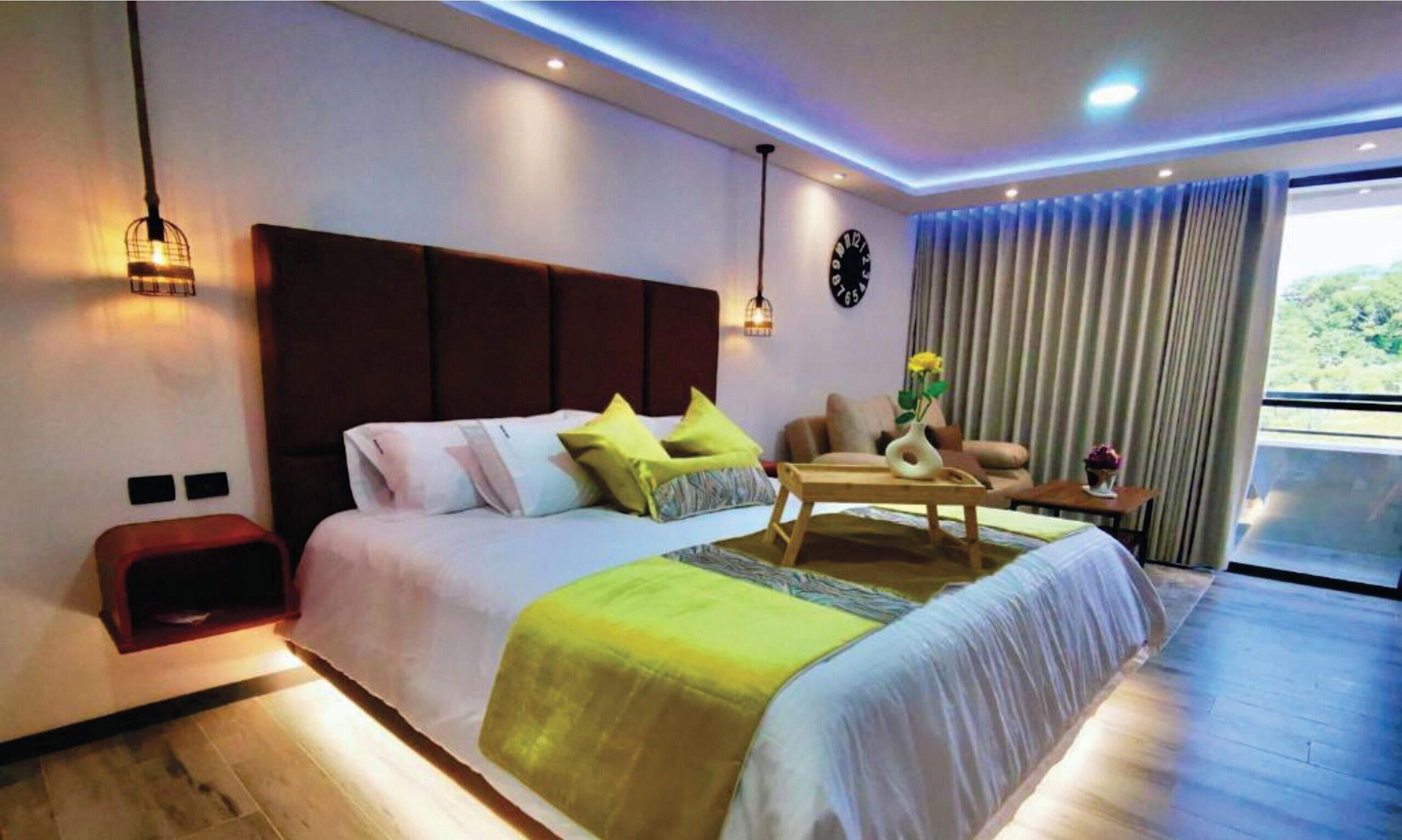 A modern hotel room with a large bed, a wooden side table, a sofa, and a view of the outdoors.