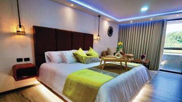 A modern hotel room with a large bed, a wooden side table, a sofa, and a view of the outdoors.