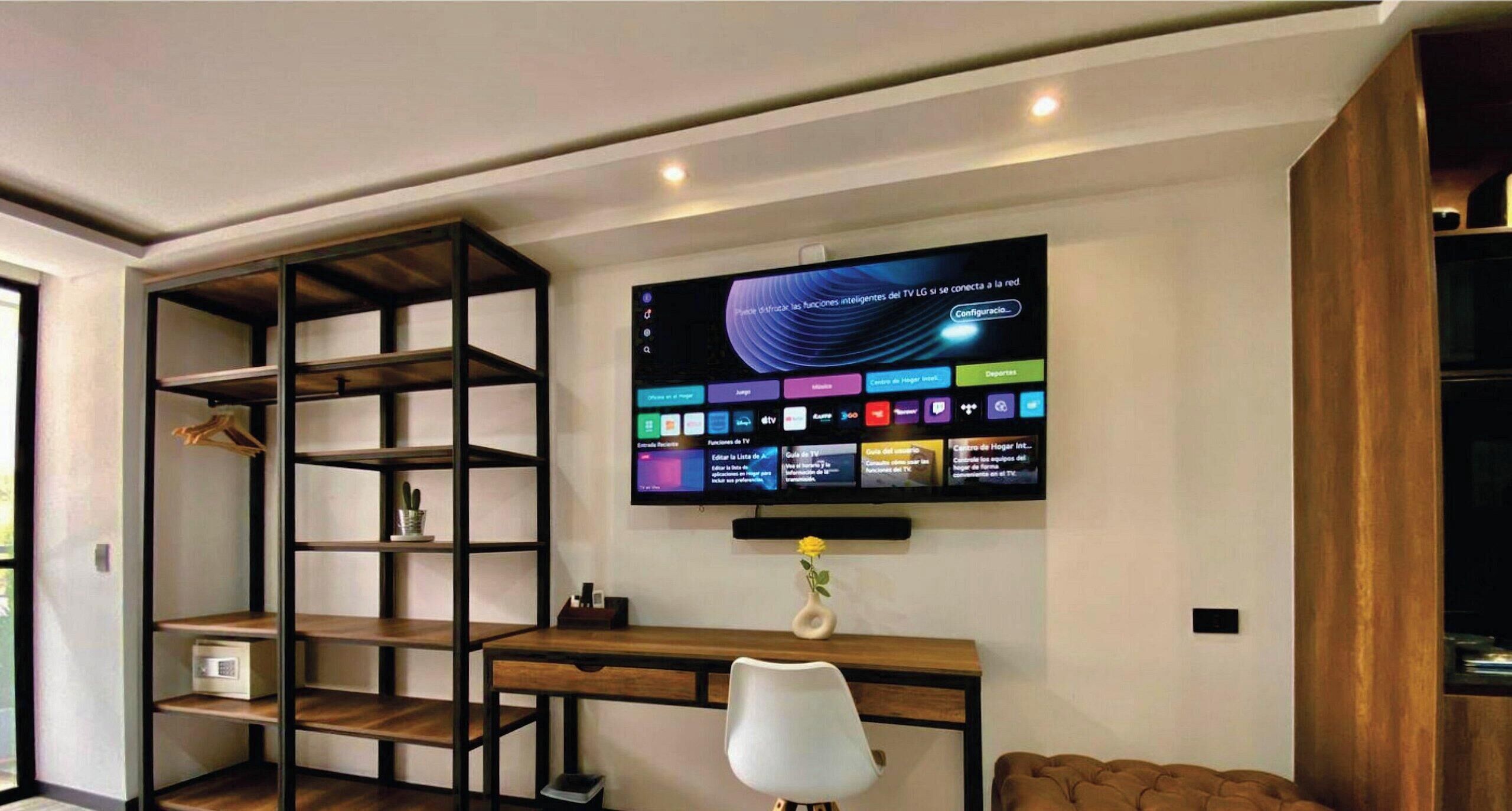 Un saló modern amb televisió plana penjada a la paret, escriptori de fusta, cadira blanca i sofà marró.