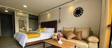 A modern hotel room with a large bed, a sofa, a wooden coffee table, and a wall clock.