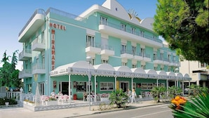Ein mehrstöckiges Hotel mit grüner Fassade und weißen Balkonen. Im Erdgeschoss befinden sich Sitzgelegenheiten im Freien und eine Bar namens 'BAR MIRAMARE'.